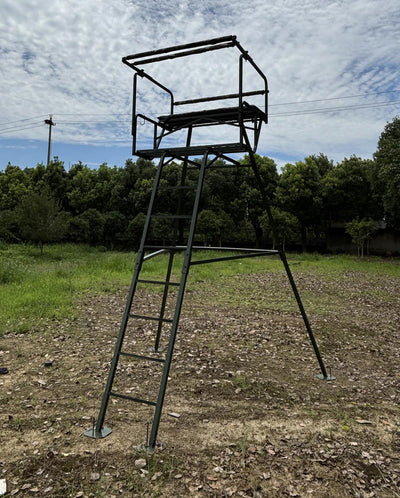 A free-standing metal hunting stand with a platform and ladder, designed for two persons, set in an outdoor setting with trees in the background.
